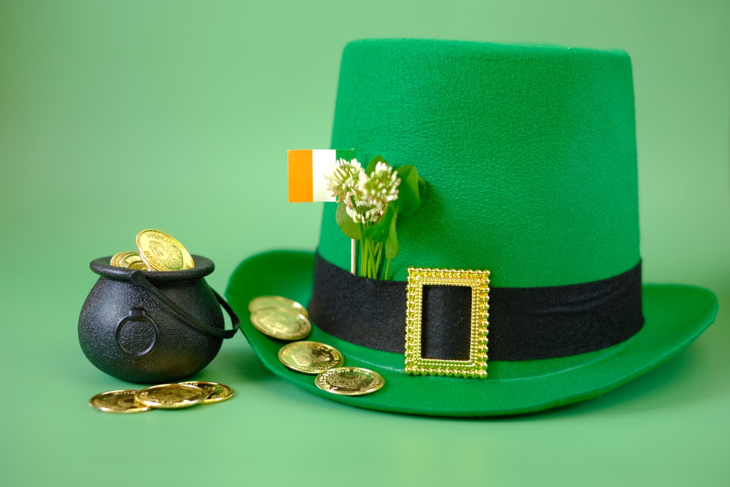 A green leprechaun hat with blooming clovers and an Irish flag in its band, next to a miniature pot of gold