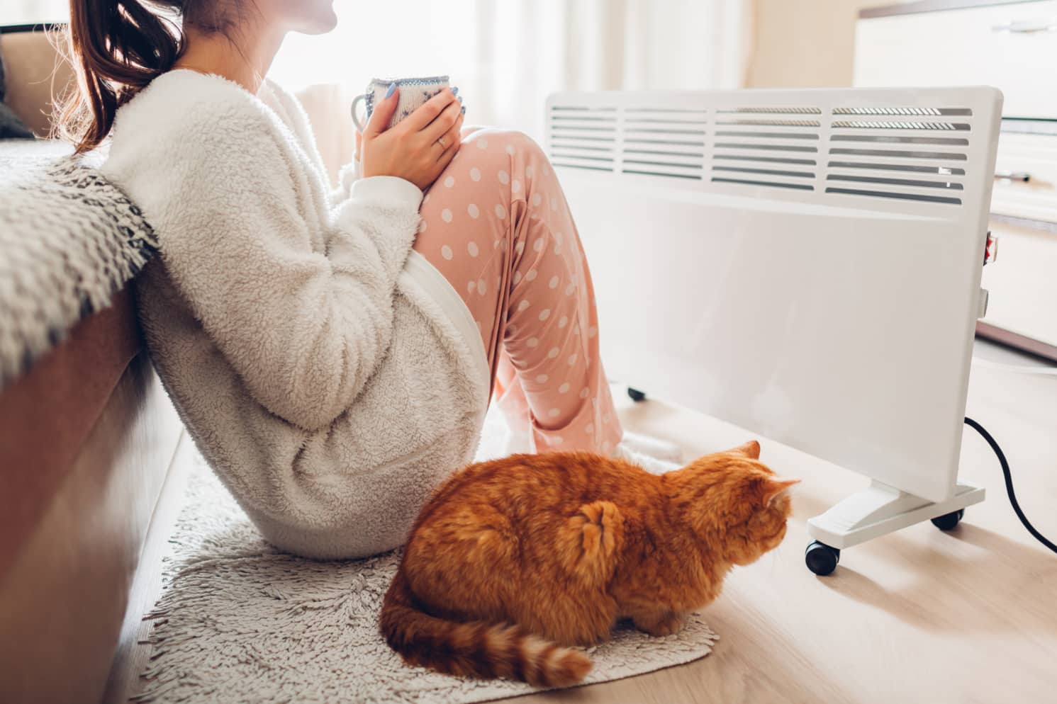 Woman with her cat sitting next to an electric heater, wearing warm clothes and drinking tea