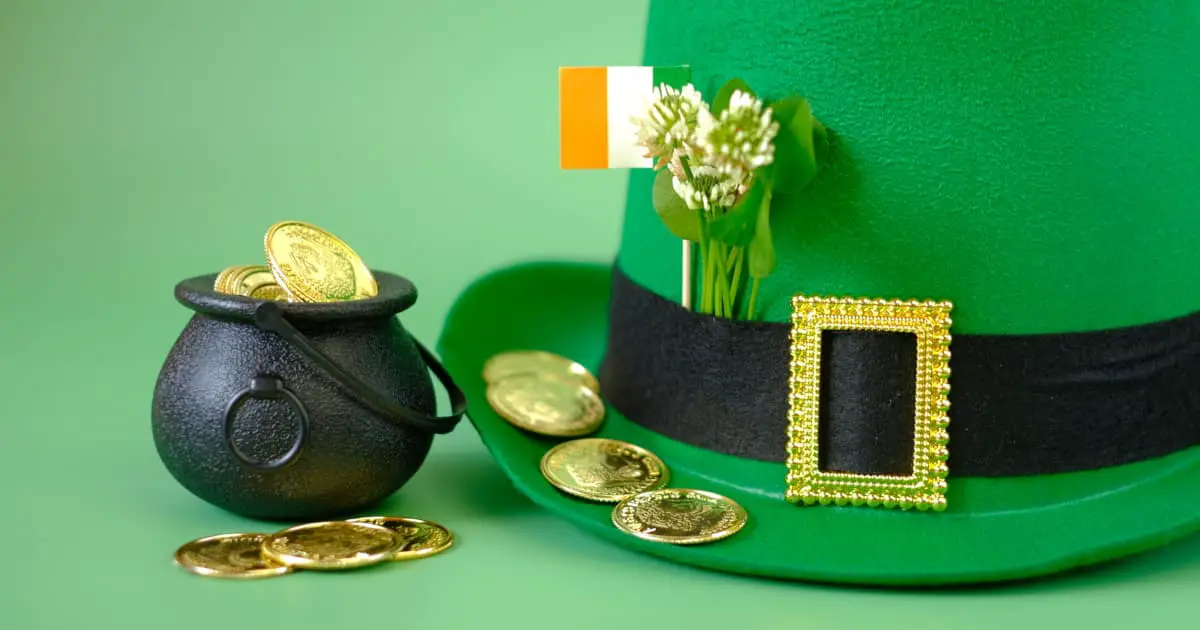A green leprechaun hat with blooming clovers and an Irish flag in its band, next to a miniature pot of gold